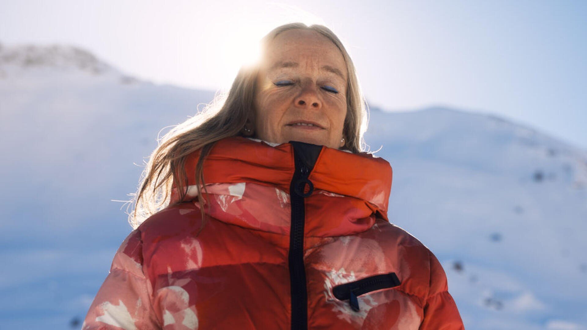 Eine Person in einer roten Winterjacke mit Blumenmuster steht draußen mit geschlossenen Augen, während das Sonnenlicht hinter ihrem Kopf scheint und im Hintergrund schneebedeckte Berge zu sehen sind.