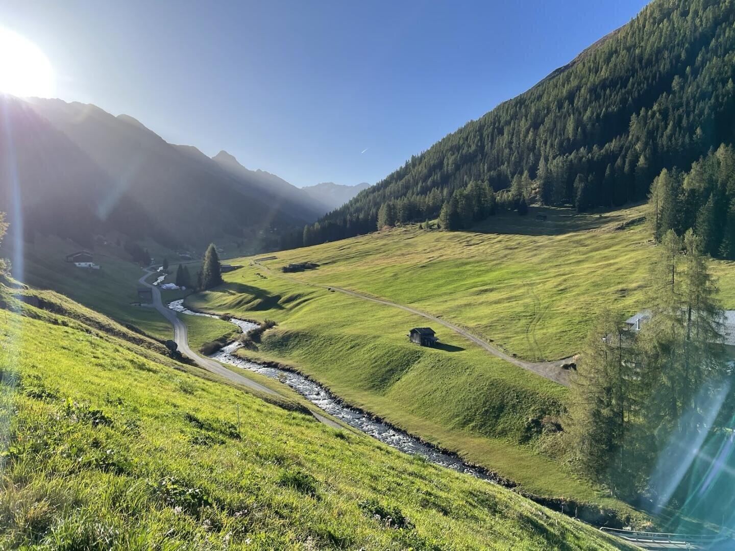 Ein sonnenbeschienenes Tal mit grünen Grashügeln, einem gewundenen Bach, verstreuten Hütten und bewaldeten Bergen im Hintergrund unter einem klaren blauen Himmel.