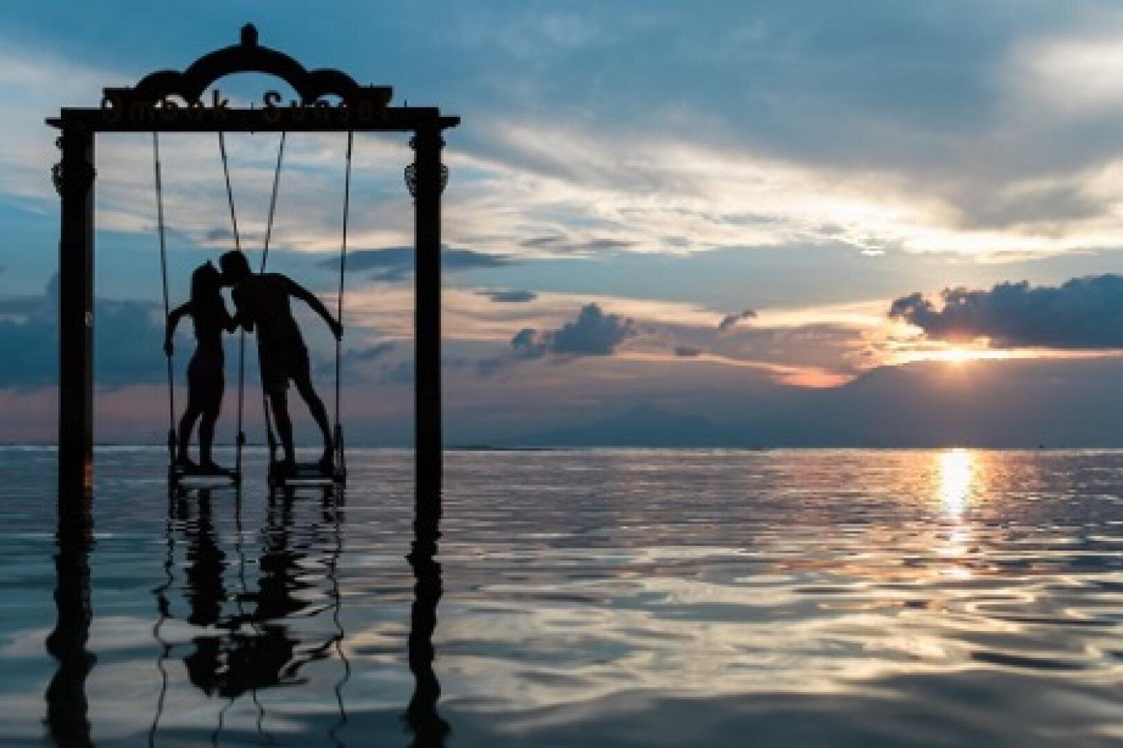 Zwei Menschen stehen bei Sonnenuntergang auf Schaukeln über dem Wasser; ihre Silhouetten zeichnen sich ab, während sie sich unter einem verzierten Rahmen zueinander beugen, um sich zu küssen, vor dem Hintergrund dramatischer Wolken und Berge.