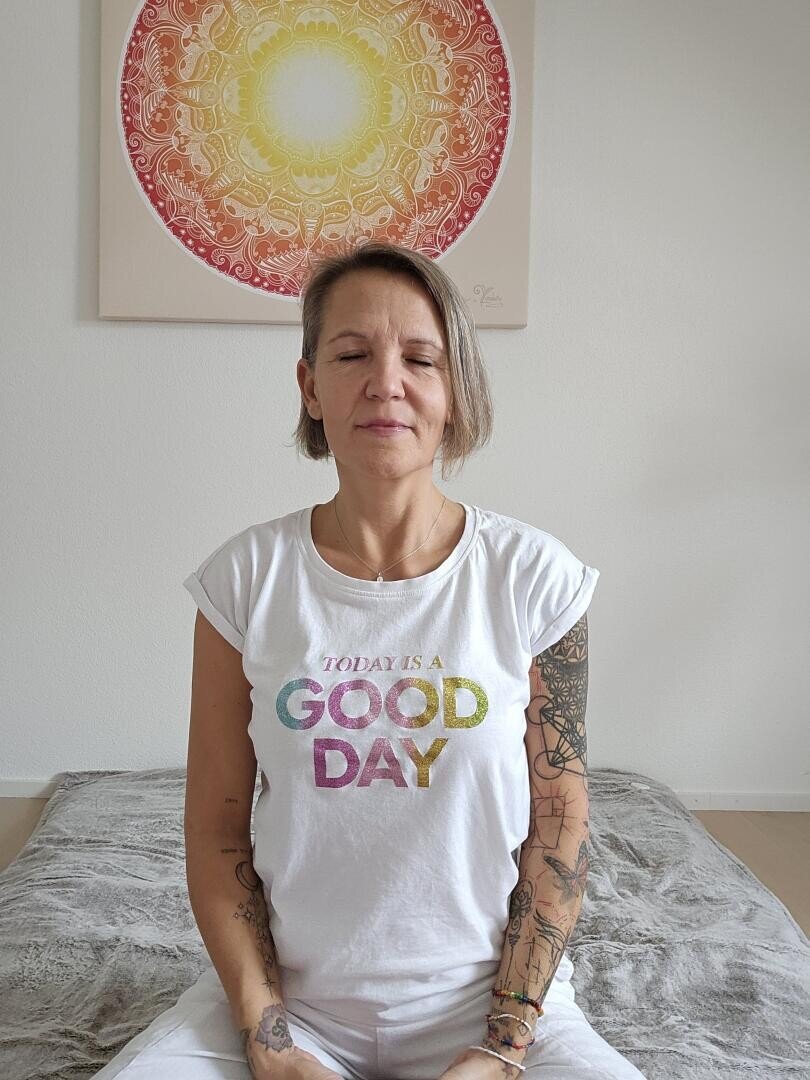 Eine Frau mit kurzen Haaren und Tattoos sitzt im Schneidersitz auf einer Matte, die Augen geschlossen und entspannt, und trägt ein weißes T-Shirt mit der Aufschrift TODAY IS A GOOD DAY. An der Wand hinter ihr hängt ein buntes Mandala-Kunstwerk.