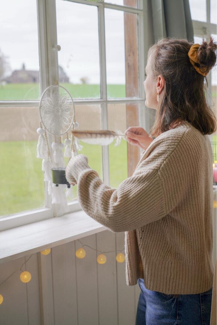 Eine Frau in einem beigen Pullover steht am Fenster, in der Hand hält sie eine Feder und einen weißen Traumfänger; auf der Fensterbank liegen Lichterketten, und draußen ist ein grünes Feld zu sehen.