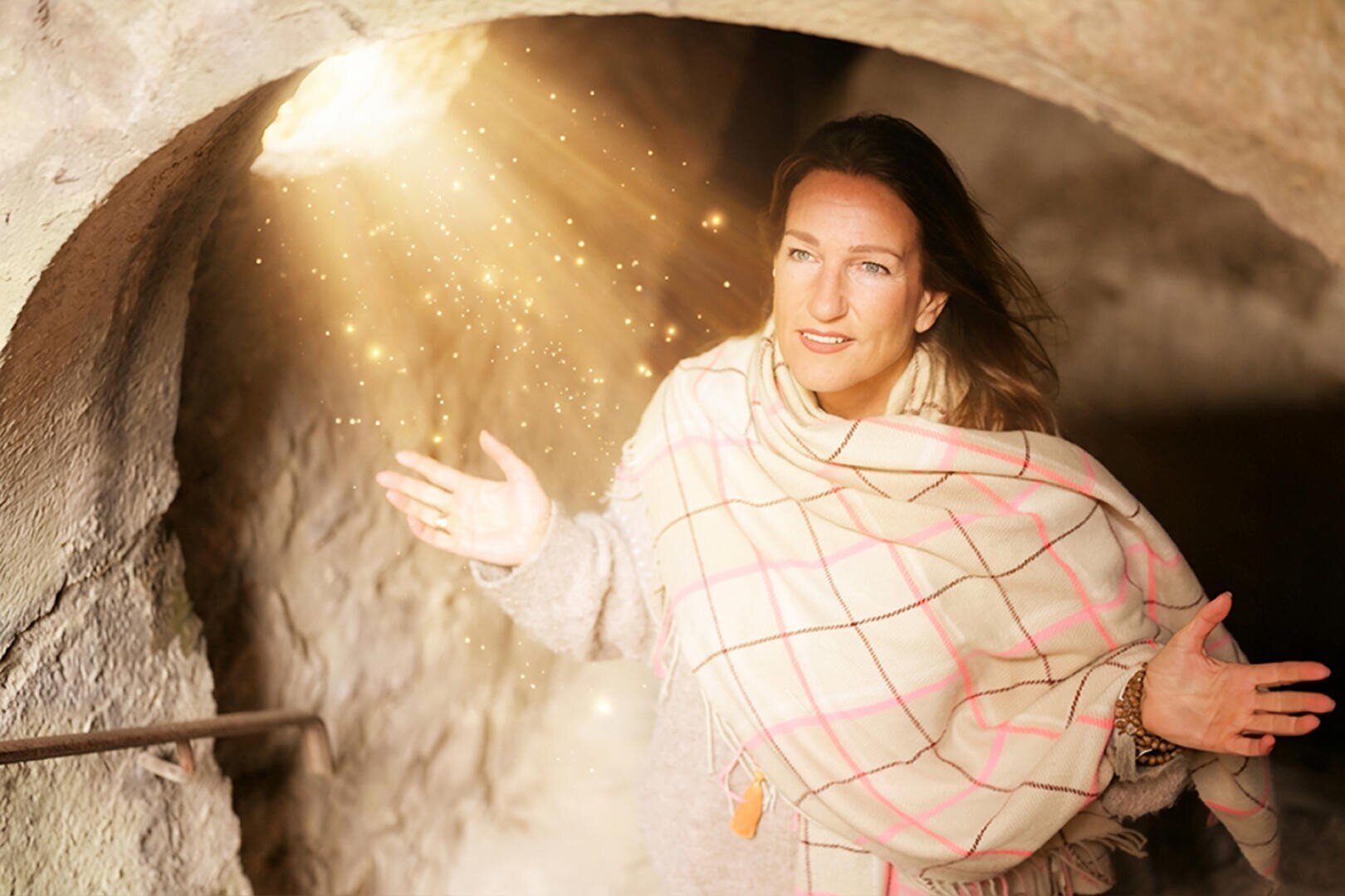 Eine Frau, die einen karierten Schal trägt, steht in einem Steintunnel, der von einem warmen, magischen Licht erhellt wird, in dem goldene Funken von oben herabschimmern. Sie hält ihre Arme offen und lächelt sanft in die Kamera.