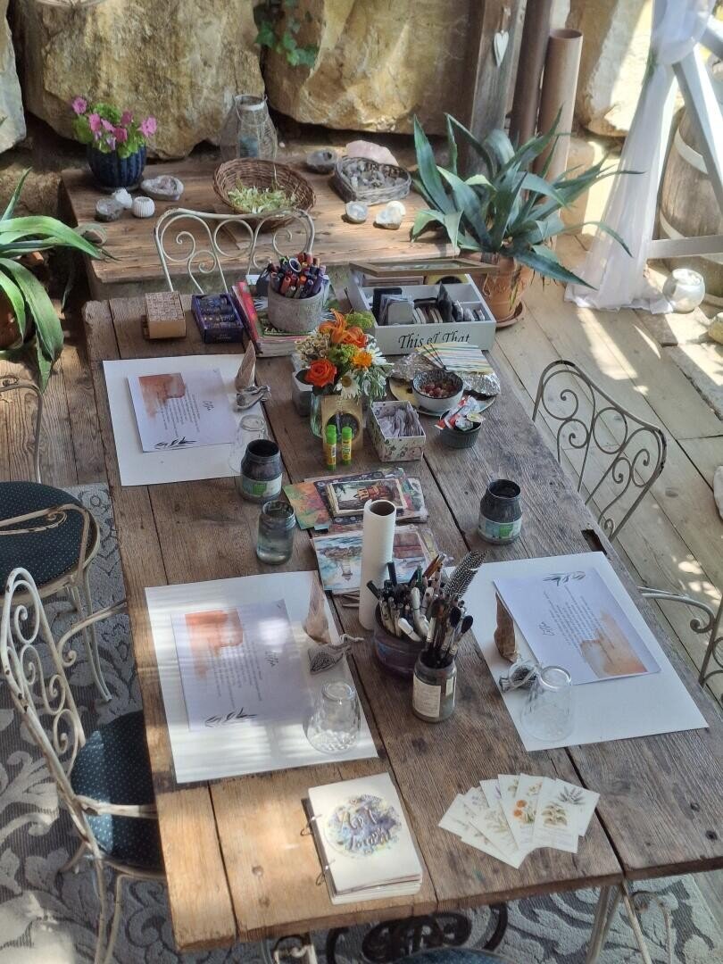 Ein rustikaler Holztisch, gedeckt zum Malen, mit Malutensilien, Aquarellpaletten, Pinseln, Papier, Blumen und Pflanzen in einem sonnendurchfluteten Raum mit Steinwänden und Metallstühlen.