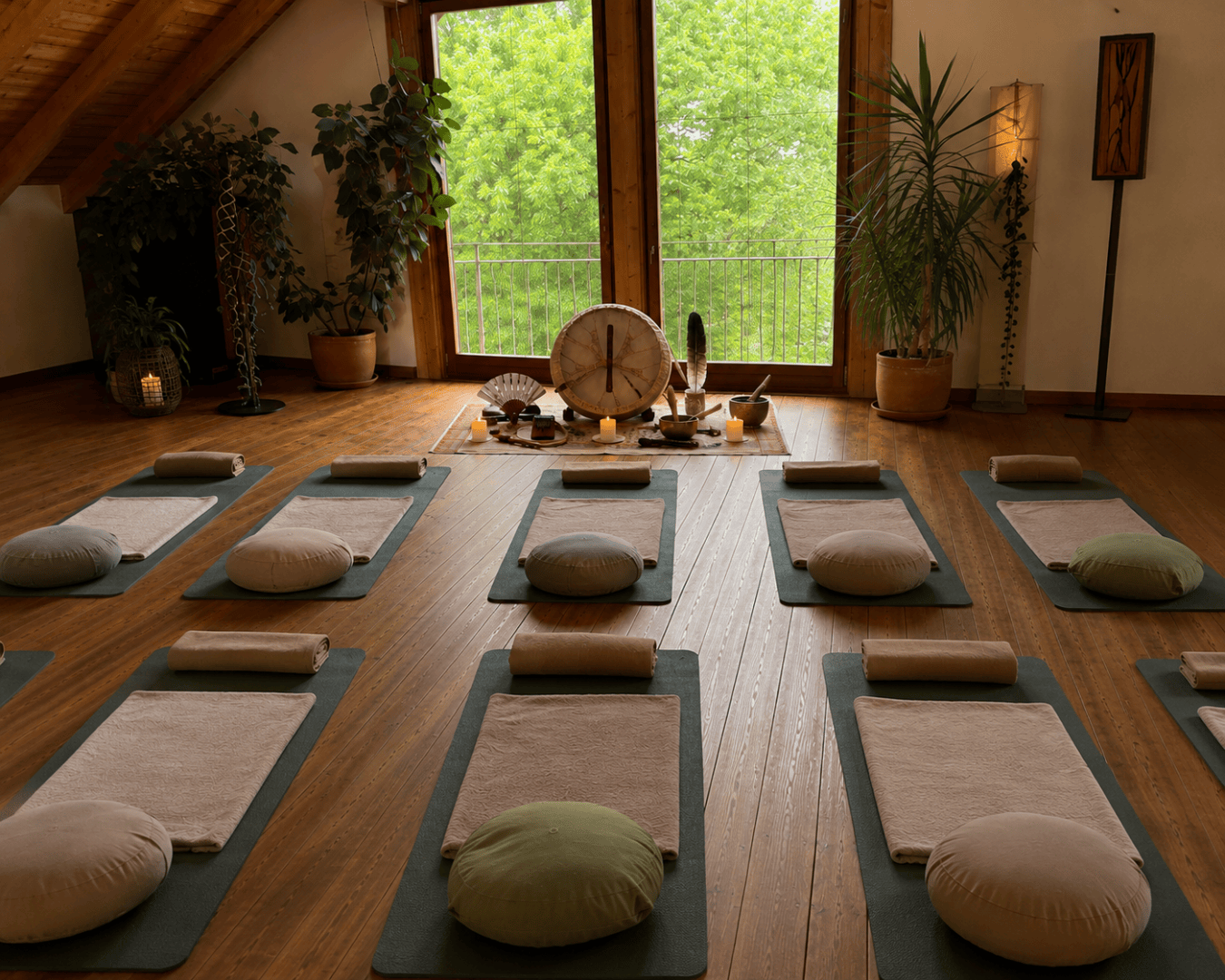 Ein ruhiges Yoga-Studio, in dem Matten, Kissen und Decken ordentlich auf einem Holzboden ausgelegt sind. Durch große Fenster bietet sich ein Blick auf die üppige grüne Landschaft draußen. Im vorderen Teil des Raums stehen Pflanzen und ein dekorativer Altar.