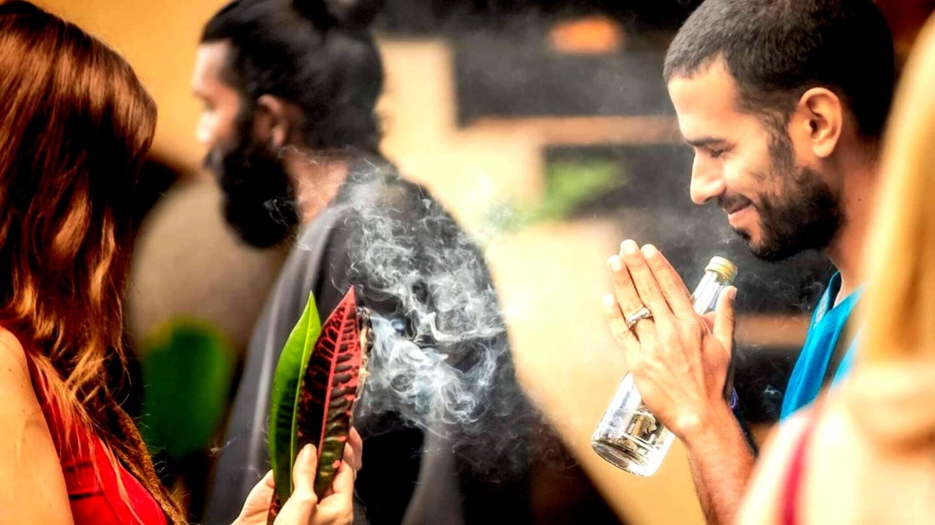 Ein Mann lächelt mit zum Gebet erhobenen Händen, hält eine Flasche in der Hand und blickt auf eine Frau, die ein buntes Blatt und Rauch in der Hand hält. Eine weitere Person mit Bart ist unscharf im Hintergrund zu erkennen. Die Stimmung ist ruhig und feierlich.