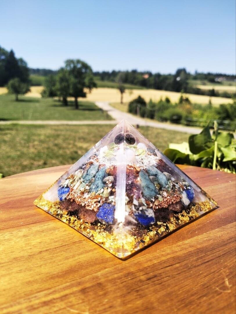 Eine dekorative Pyramide aus Kunstharz mit eingearbeiteten Kristallen steht im Freien auf einer Holzunterlage, im Hintergrund sind eine grasbewachsene Landschaft, Bäume und ein strahlend blauer Himmel zu sehen.