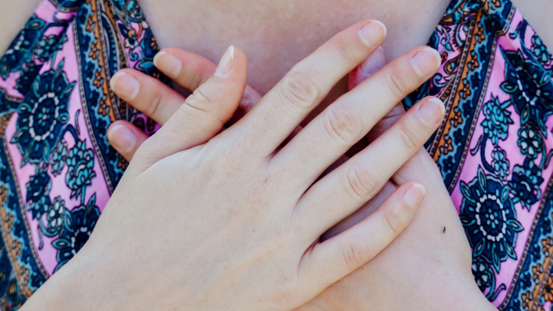 Nahaufnahme der über der Brust gekreuzten Hände einer Person, die Finger leicht gespreizt. Der Hintergrund zeigt einen Teil eines gemusterten Kleidungsstücks und nackte Haut. Das Bild vermittelt ein Gefühl der Ruhe, des Komforts oder der Selbstfürsorge.