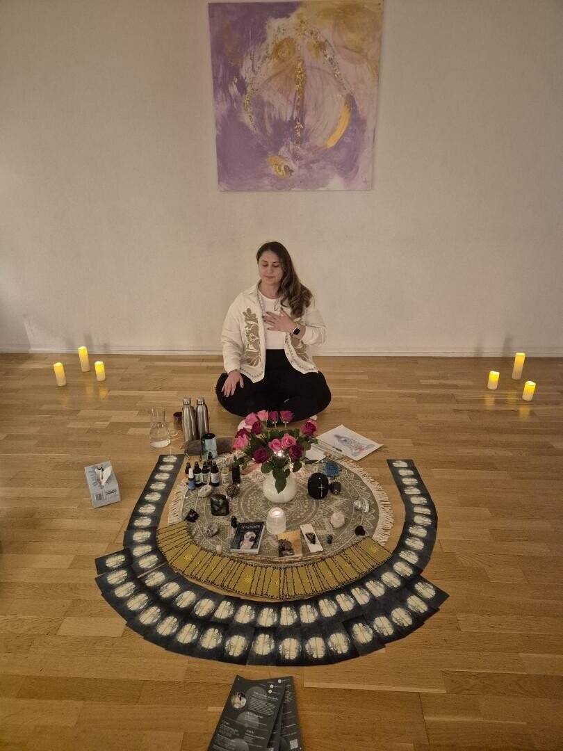 Eine Frau sitzt im Schneidersitz auf einem Holzboden, die Augen geschlossen, die Hand auf der Brust, umgeben von Kerzen, Blumen, Ritualgegenständen, Karten und Büchern. An der Wand hinter ihr hängt ein lila abstraktes Gemälde.