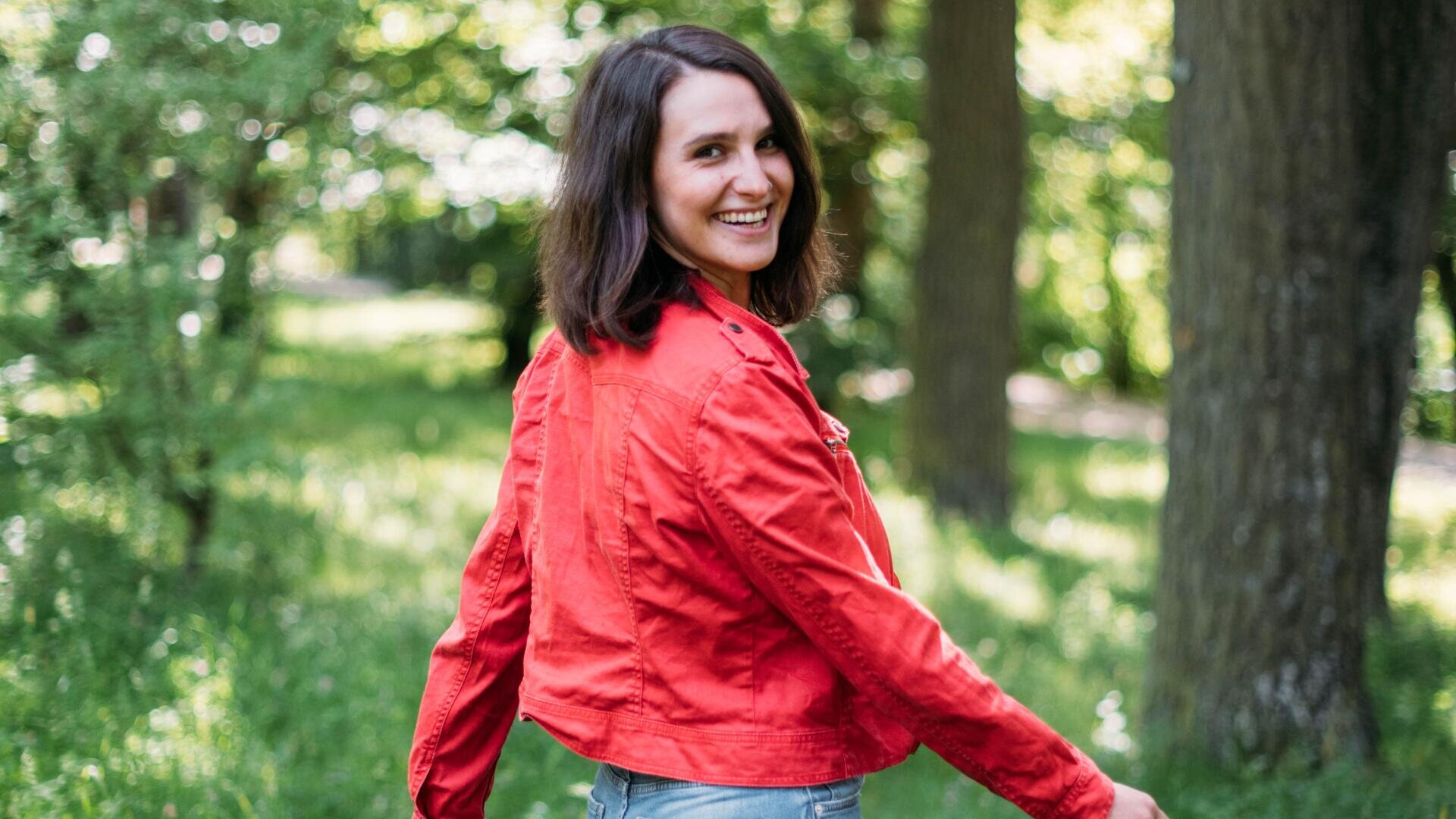 Eine Frau in einer roten Jacke lächelt, als sie sich umdreht, während sie durch einen sonnendurchfluteten, grünen Wald mit hohen Bäumen im Hintergrund geht.