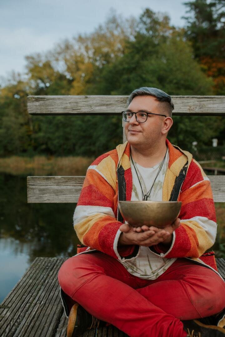 Eine Person mit Brille und einem gestreiften Kapuzenpulli sitzt auf einem Holzsteg und hält eine Klangschale in der Hand.