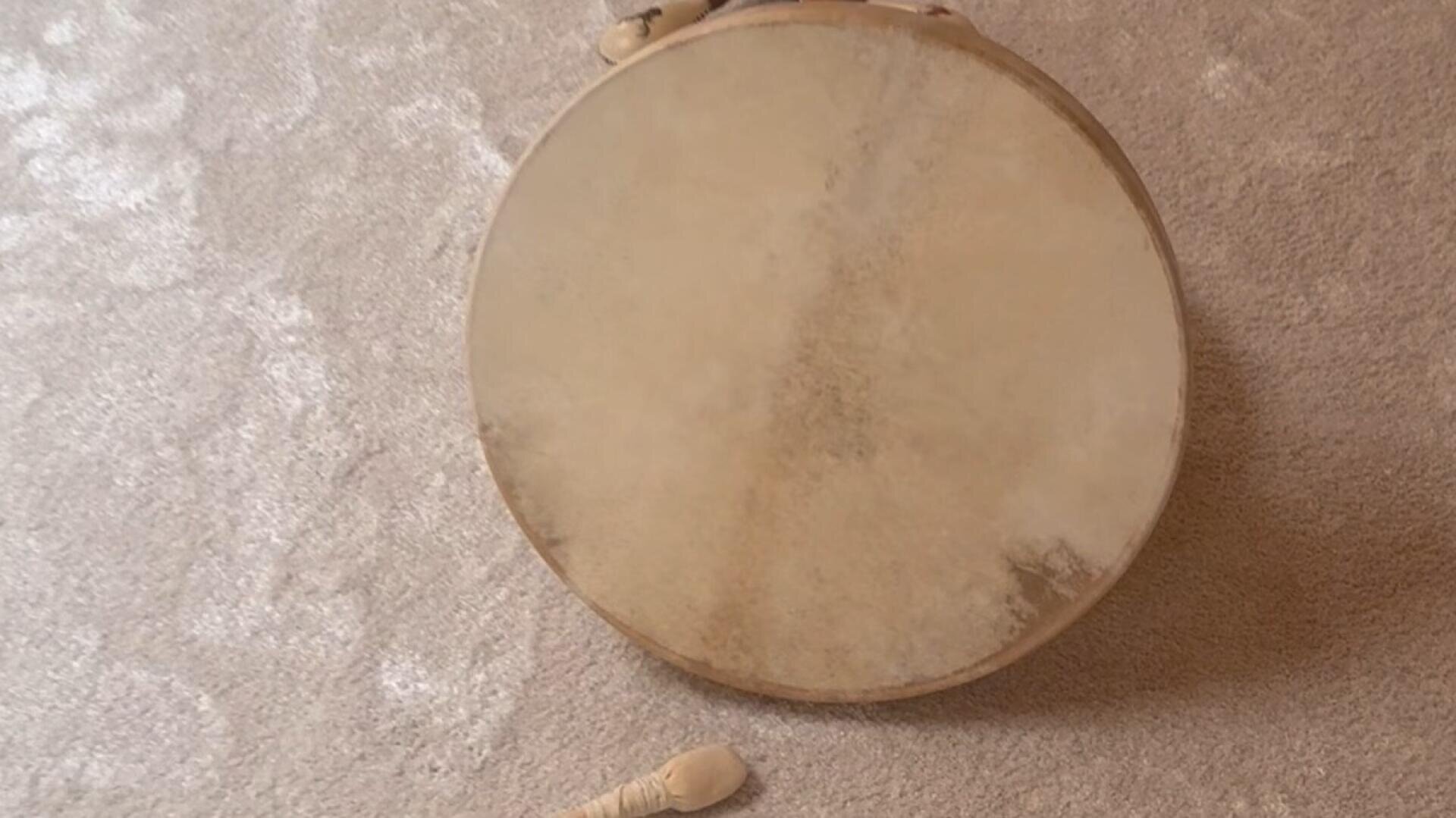 Eine runde, handliche Trommel mit einer rustikalen, naturbelassenen Felloberfläche steht auf einem hellen Teppich. Daneben liegt ein Trommelstock aus Holz.