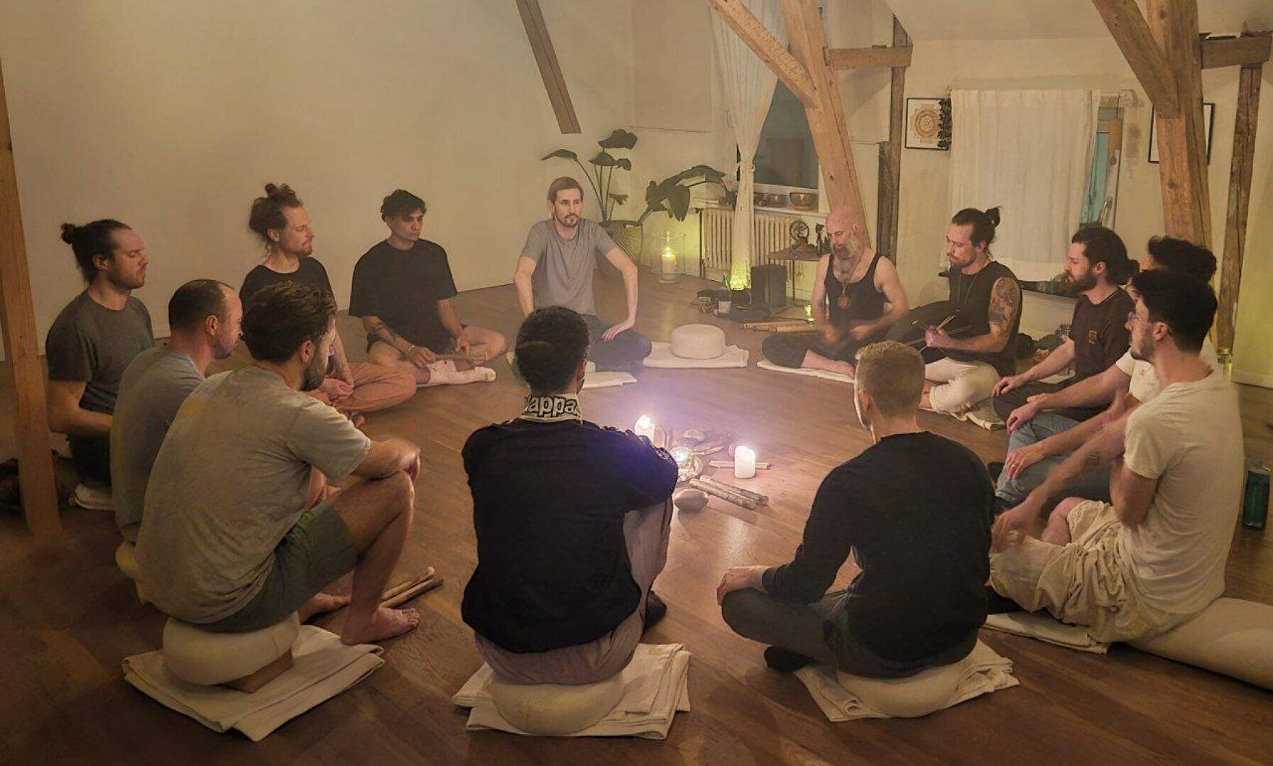 Eine Gruppe von Menschen sitzt in einem Kreis auf Kissen in einem sanft beleuchteten Raum, mit Kerzen und Gegenständen in der Mitte, und scheint zu meditieren oder an einem Gruppenritual teilzunehmen. Im Hintergrund sind Holzbalken und Zimmerpflanzen zu sehen.