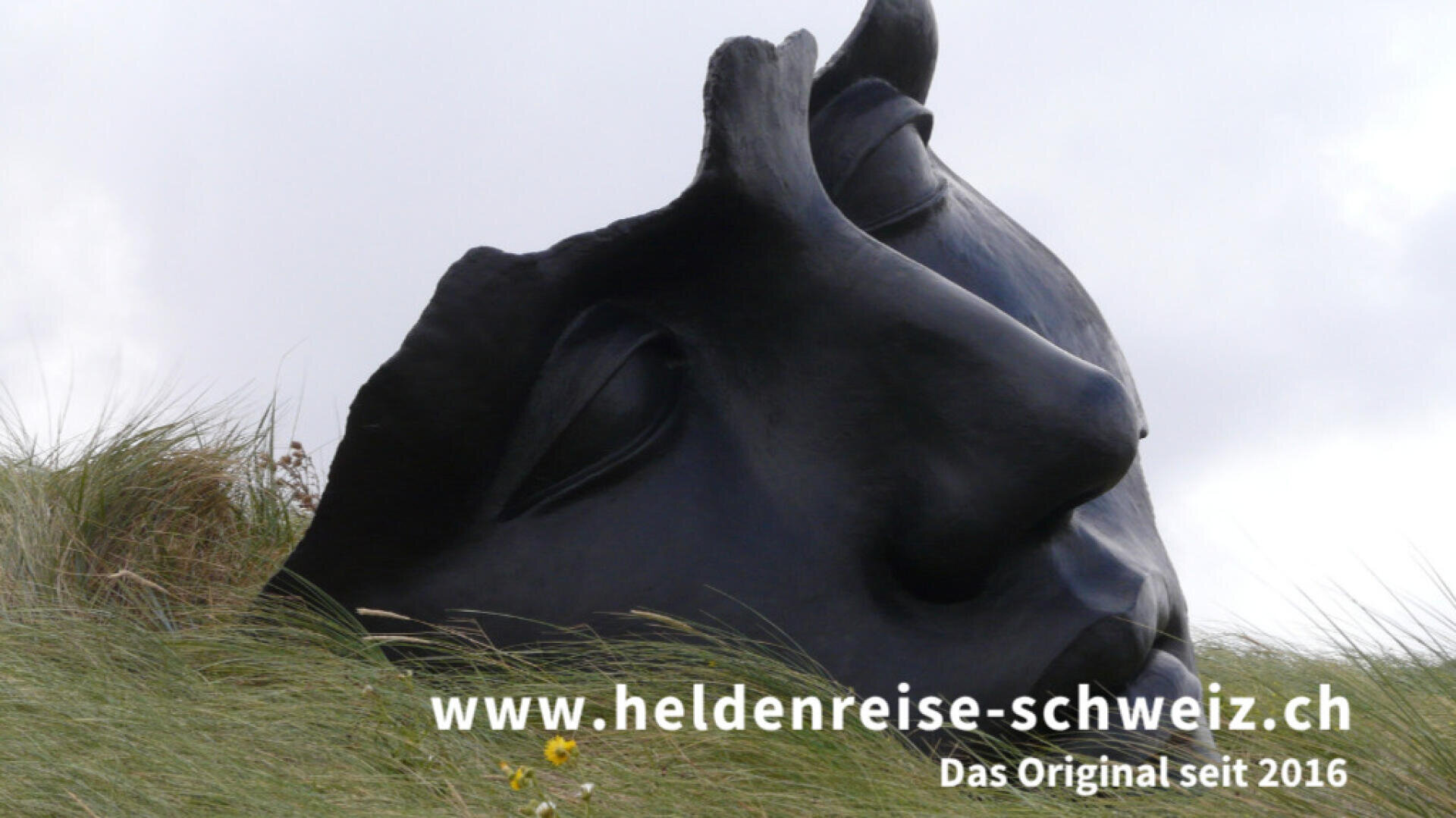 Eine große, schwarze Skulptur eines menschlichen Gesichts liegt auf der Seite im hohen Gras. Der Text am unteren Rand lautet: www.heldenreise-schweiz.ch Das Original seit 2016.