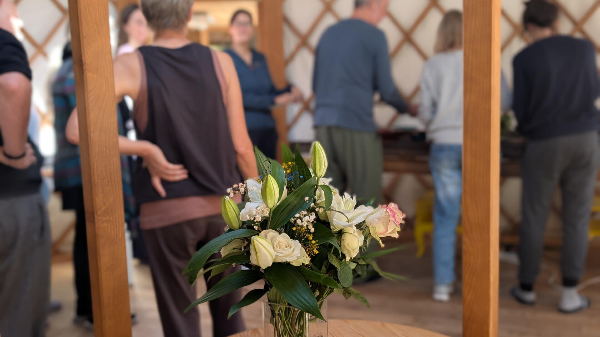 Eine Vase mit weißen und zartrosa Blumen und Grünzeug steht auf einem runden Tisch neben zwei kleinen Zierkürbissen, während sich im Hintergrund eine Gruppe von Menschen in einer Holzjurte unterhält.