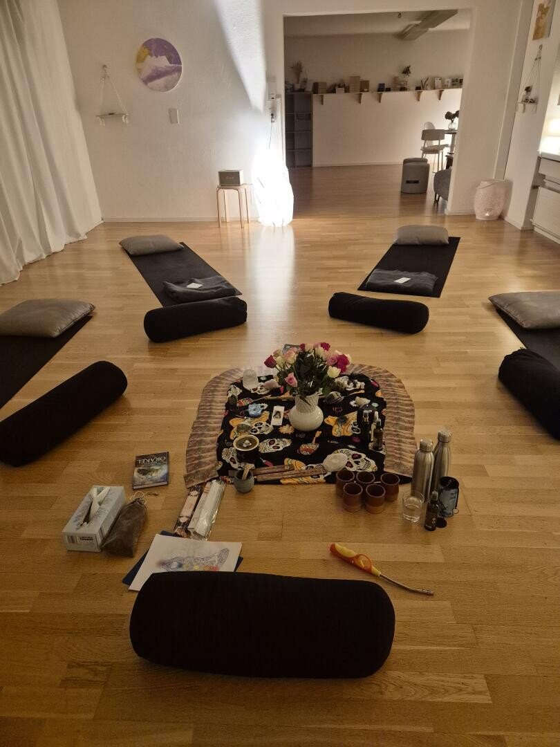 Ein gemütlicher Raum mit fünf Bodenmatten und Kissen, die kreisförmig um einen dekorativen Mittelteil mit Blumen, Kerzen und verschiedenen Gegenständen angeordnet sind. Sanftes Licht und eine warme, einladende Atmosphäre erfüllen den Raum mit Holzfußboden.