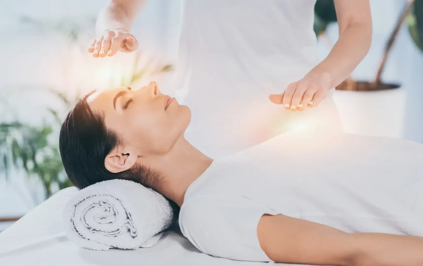 Eine Frau liegt mit geschlossenen Augen auf einer Massageliege und stützt ihren Kopf auf ein Handtuch, während eine andere Person ihre Hände über sie hält, mit leuchtenden Lichteffekten um die Hände, die auf Energieheilung oder Reiki-Therapie hindeuten.