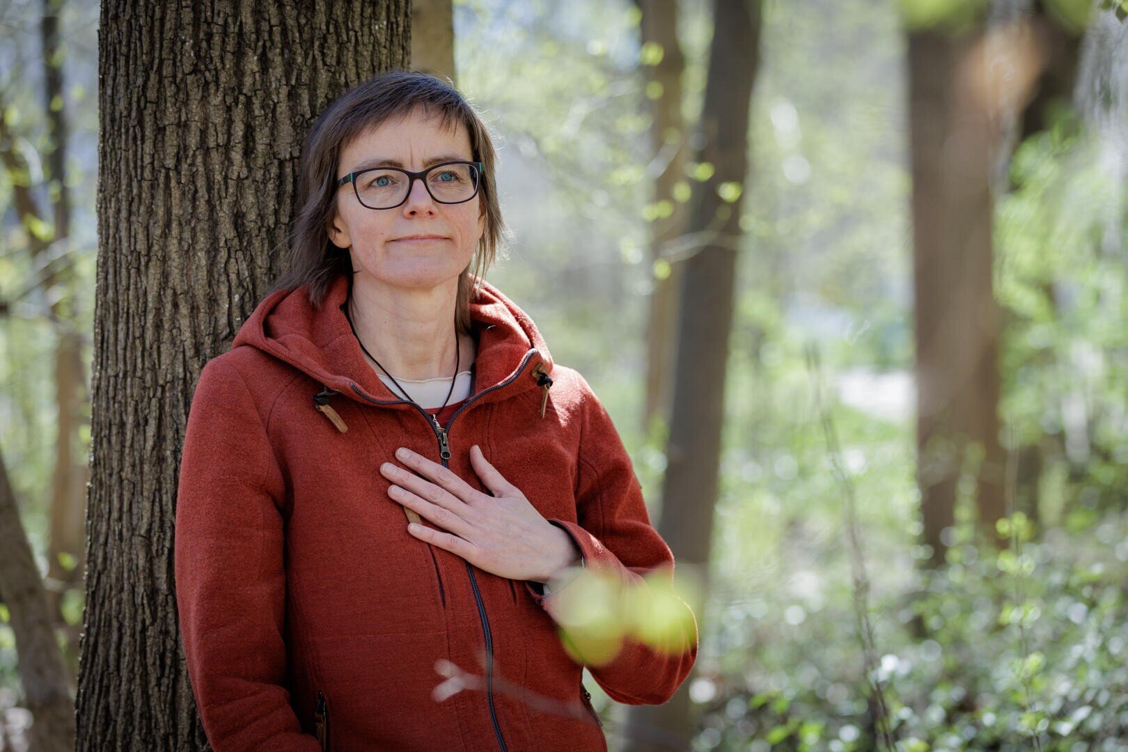 Eine Frau in einer roten Kapuzenjacke steht vor einem Baum im Wald und legt ihre Hand auf ihr Herz.