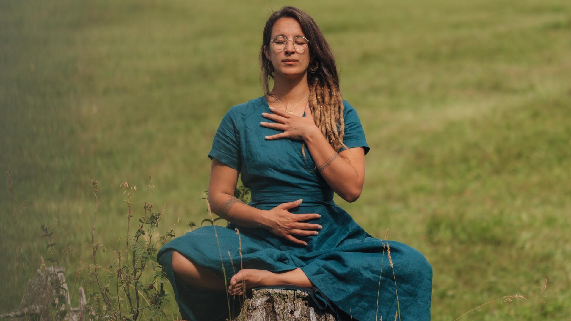 Eine Frau in einem blaugrünen Kleid sitzt im Schneidersitz auf einem Baumstumpf in einer Wiese, die Augen geschlossen, eine Hand auf der Brust, die andere auf dem Bauch, sie wirkt ruhig und meditativ.