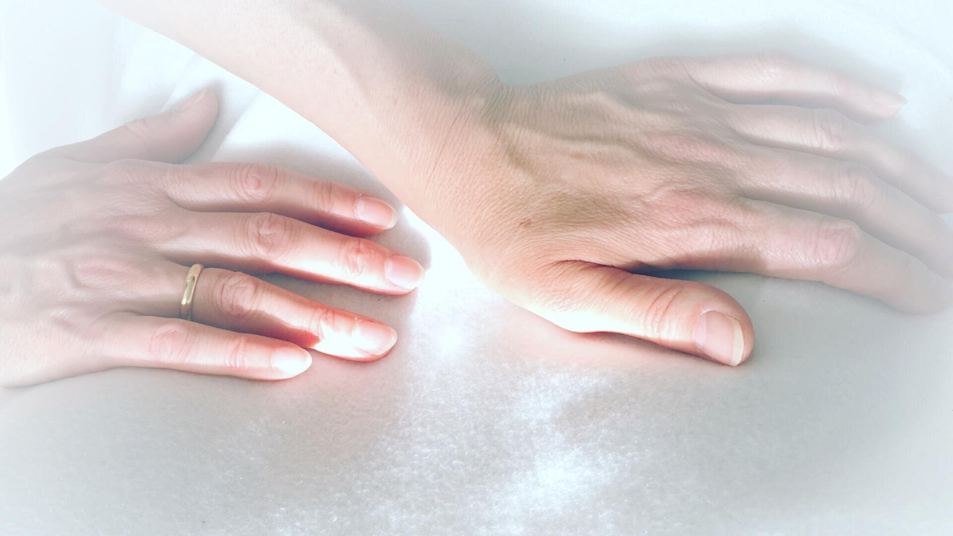 Zwei Hände ruhen sanft auf einer weichen, weißen Oberfläche. An einer Hand befindet sich ein goldener Ring am Ringfinger. Das Bild ist sanft beleuchtet, was eine ruhige und friedliche Atmosphäre schafft.