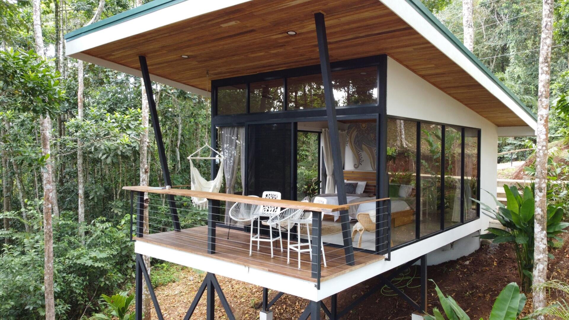 Eine moderne Hütte mit großen Glasfenstern und einem Holzdeck steht auf Stelzen in einem üppigen Wald. Die Terrasse ist mit einem weißen Tisch, Stühlen und einer Hängematte ausgestattet und von Grün umgeben.