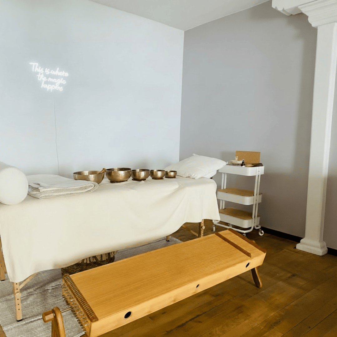 Eine Massageliege mit Klangschalen, eine Bank und ein kleiner Rollwagen in einem ruhigen, minimalistisch eingerichteten Raum; an der Wand hängt eine Leuchtreklame mit der Aufschrift: „Hier geschieht das Wunder…“.