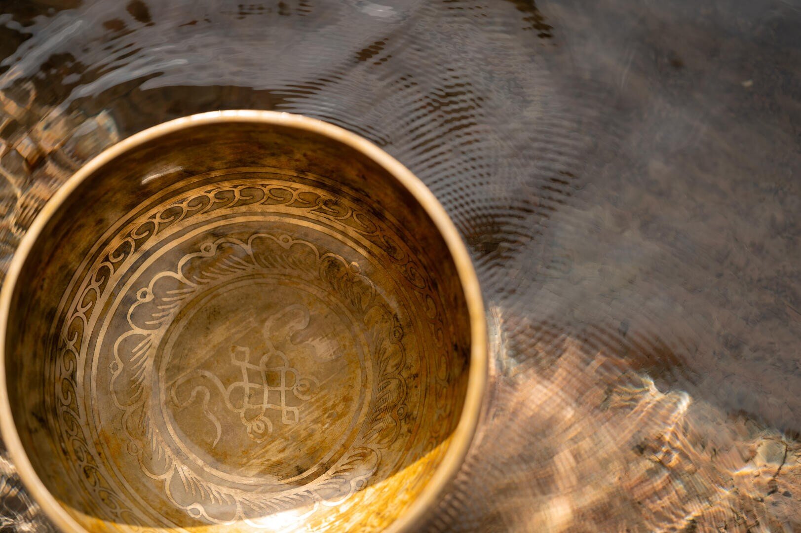 Eine Messing-Klangschale mit eingravierten Mustern liegt in plätscherndem Wasser; das Sonnenlicht spiegelt sich auf ihrer Oberfläche wider und schafft eine friedliche und beruhigende Atmosphäre.