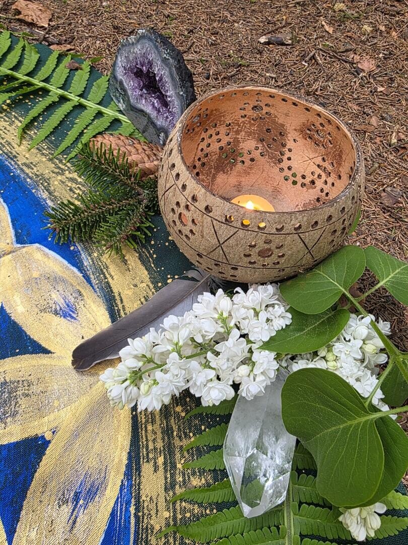 Auf einem Tuch mit gemalten Blumen steht eine dekorative Schale mit einer brennenden Kerze, umgeben von weißen Blüten, grünen Blättern, einer Feder, einem Kristall, Farnwedeln und einer Amethyst-Geode auf einem Waldboden.