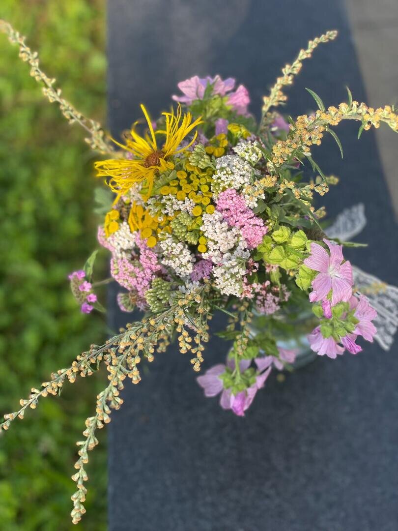 Ein Strauß Wildblumen mit gelben, rosa, weißen und violetten Blüten liegt auf einer dunklen Unterlage, im Hintergrund ist Grün zu sehen. Die Blumen sind in Form und Größe unterschiedlich und bilden ein natürliches, rustikales Arrangement.