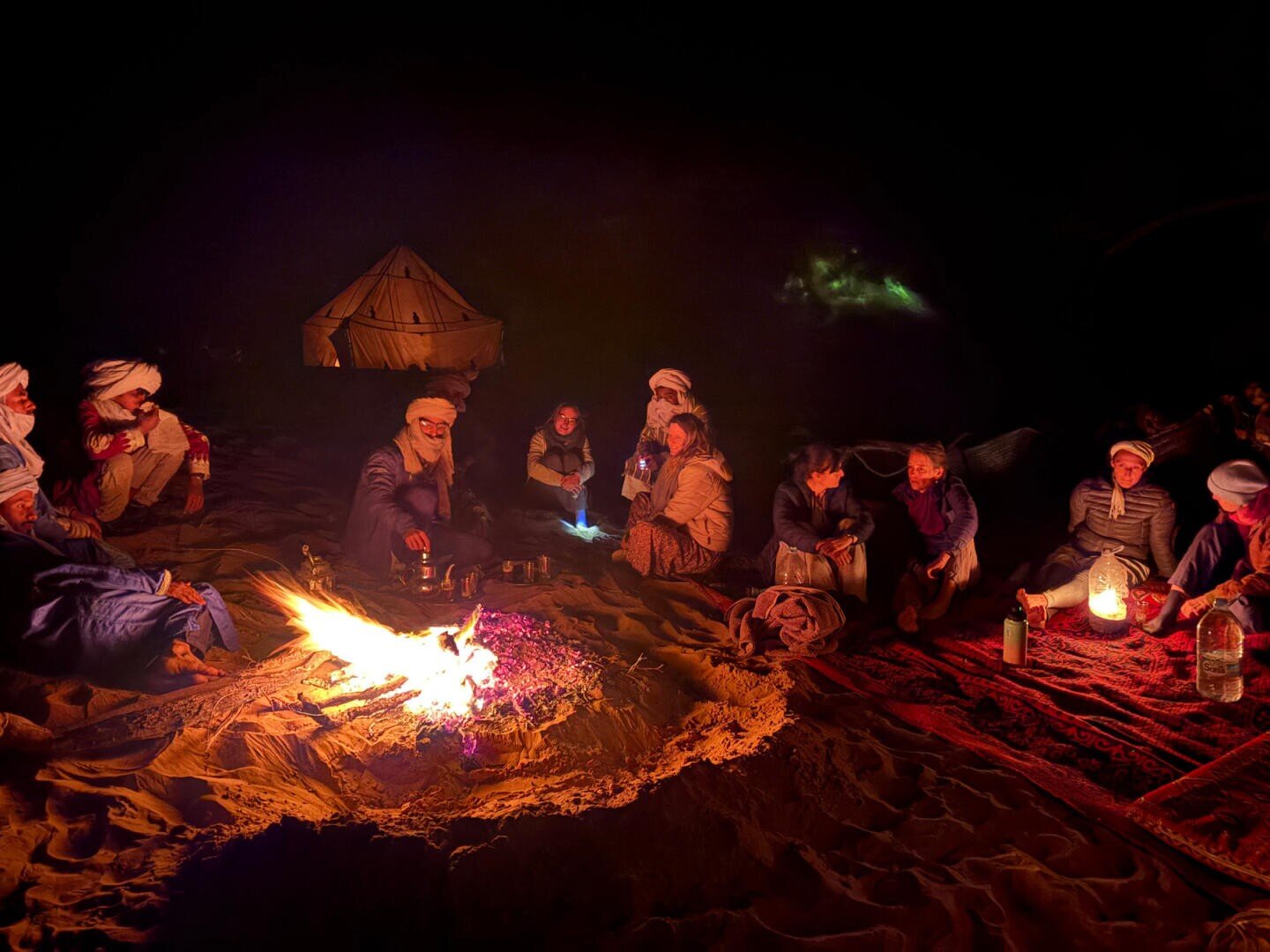 Eine Gruppe von Menschen mit Kopftüchern sitzt nachts in der Wüste um ein Lagerfeuer herum; im Hintergrund ist ein Zelt zu sehen, und auf dem Sand sowie auf einem Teppich in der Nähe stehen verschiedene Behälter.