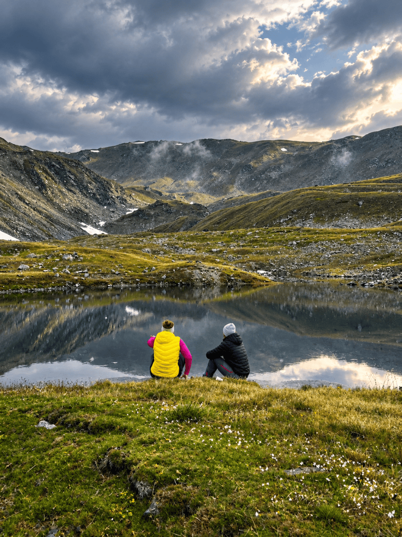 Zwei Menschen sitzen an einem ruhigen Bergsee, umgeben von felsigen Hügeln und grasbewachsenen Hängen unter einem stimmungsvollen, bewölkten Himmel, und genießen die malerische Landschaft.