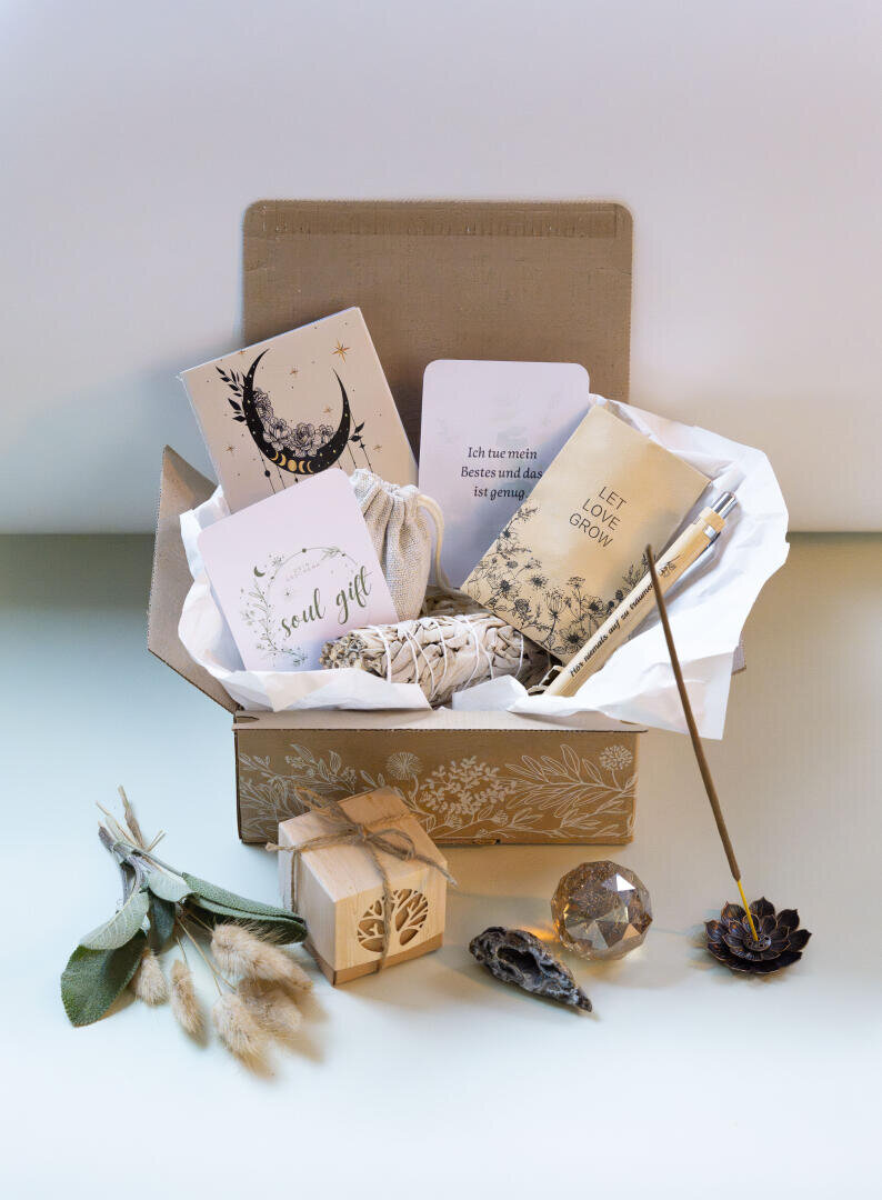 Eine Geschenkbox, gefüllt mit Gegenständen für die Selbstfürsorge, darunter ein Notizbuch, Karten, Räucherstäbchen, ein kleines Handtuch, ein Bündel Salbei, ein Kristall, getrocknete Blumen und ein Holzkästchen, alles fein säuberlich auf einer hellen Fläche angeordnet.
