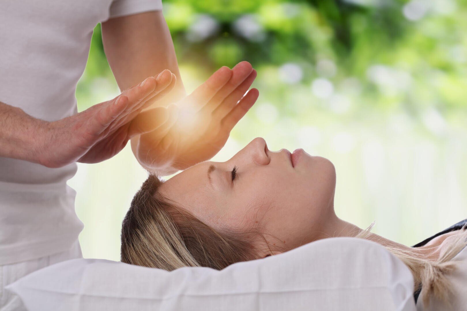 Eine Person liegt mit geschlossenen Augen, während eine andere Person ihre Hände über die Stirn hält und ein sanftes Licht ausstrahlt, das auf eine Heilungs- oder Energietherapie-Sitzung hindeutet, mit einem grünen, verschwommenen Hintergrund.