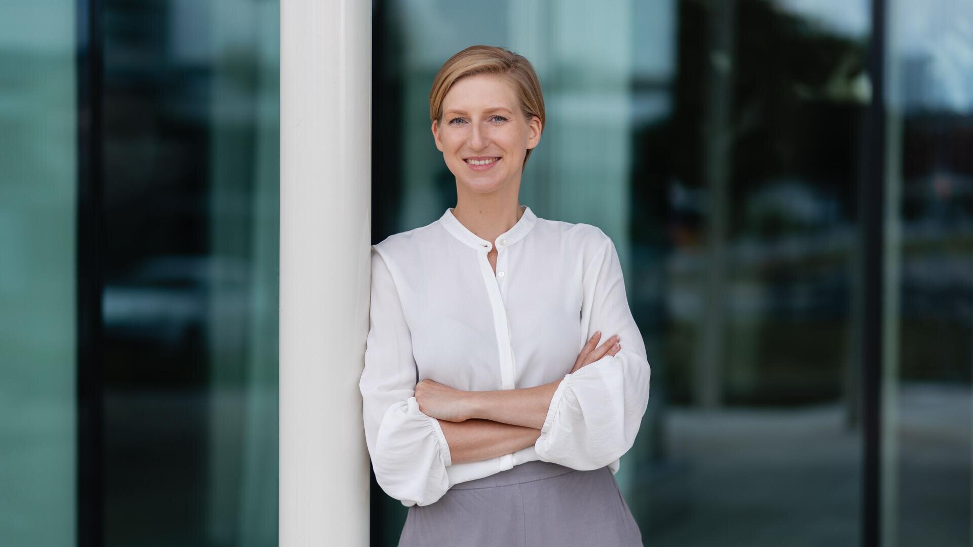 Eine Frau mit kurzen blonden Haaren, die eine weiße Bluse und eine graue Hose trägt, steht mit verschränkten Armen und lächelt vor einem modernen Glasgebäude.
