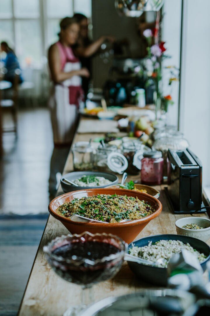 Ein Holztisch ist mit verschiedenen Gerichten gedeckt, darunter eine große Schüssel mit Salat, Beilagen und Gewürzen. Im Hintergrund bereiten zwei Personen in Schürzen das Essen in einer hellen, gemütlichen Küche zu.