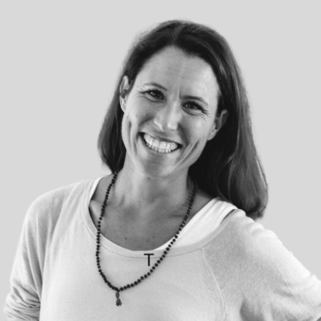 Eine Frau mit langen braunen Haaren lächelt warm, trägt ein weißes Oberteil und eine Perlenkette und steht vor einem schlichten hellgrauen Hintergrund.