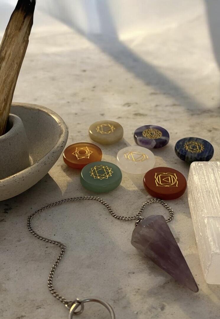 Ein Pendel, Chakra-Steine mit goldenen Symbolen, Räucherwerk und Selenit sind auf einer Marmorfläche im warmen Sonnenlicht angeordnet.