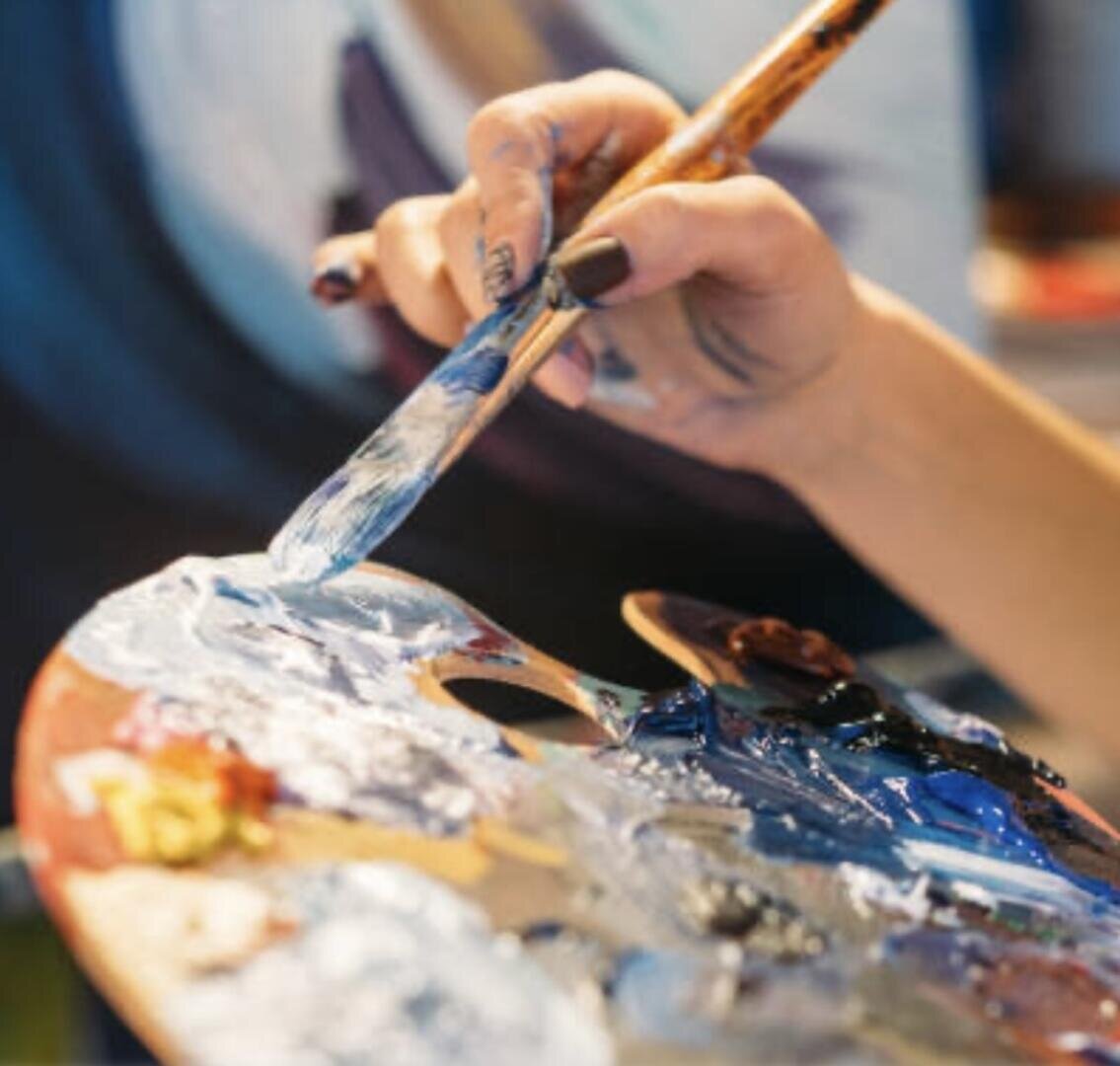 Eine Nahaufnahme einer Hand, die einen Pinsel hält und blaue und weiße Farbe auf einer Künstlerpalette mischt. Die Finger der Person haben Farbflecken, und der Hintergrund ist mit weichen Farben verschwommen.