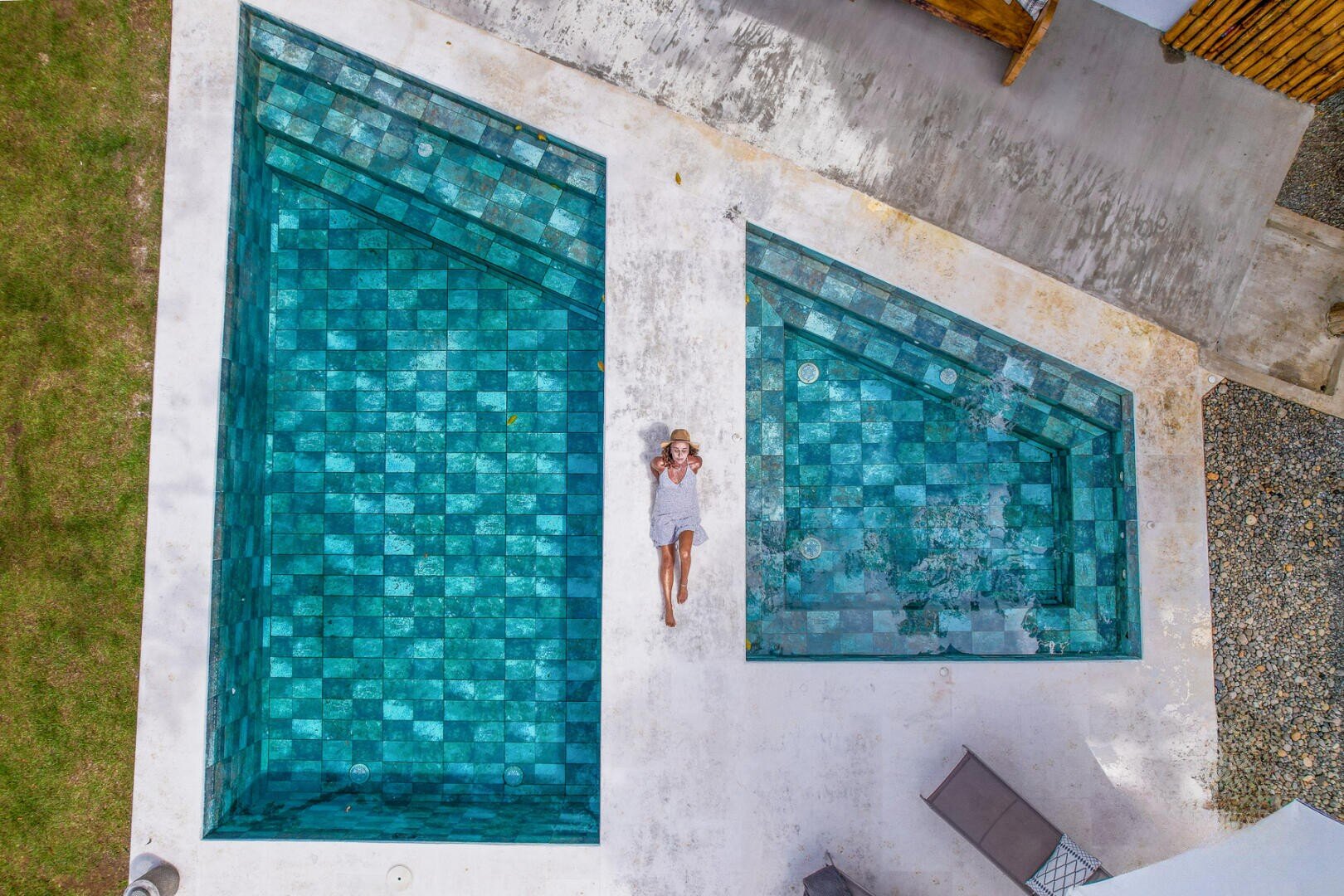 Eine Person in weißer Kleidung und mit Hut liegt auf einem weißen Pooldeck zwischen zwei blau gekachelten geometrischen Becken, von oben gesehen. An den Rändern sind Gras und Terrassenflächen zu sehen.