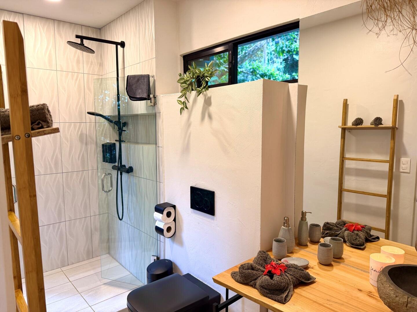 Modernes Badezimmer mit begehbarer Dusche, schwarzen Armaturen, einem Holztisch mit Handtüchern und Kerzen, Leiterregalen und einem Fenster. Die Einrichtung ist minimalistisch mit natürlichen und neutralen Tönen.