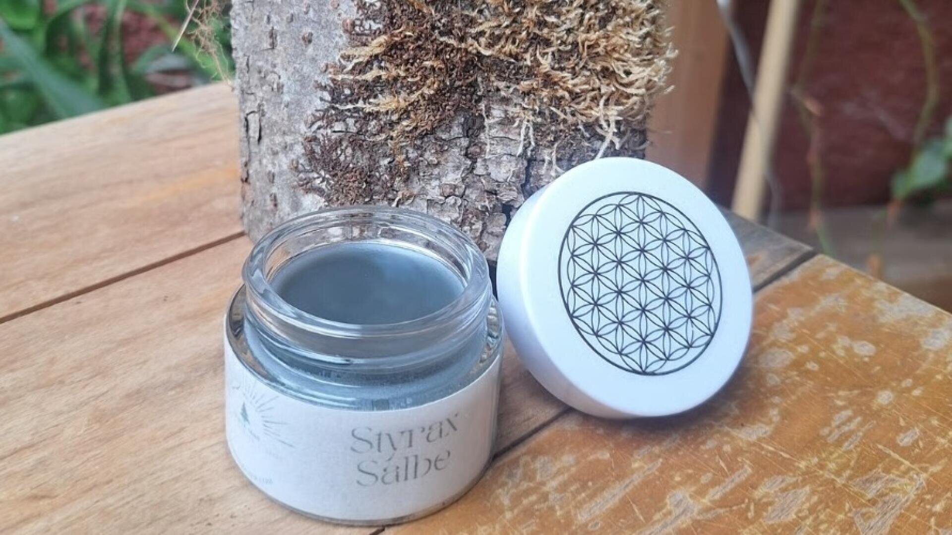 Ein Glasgefäß mit dunkler Salbe, beschriftet mit „Styrax Salbe“, steht offen auf einer Holzfläche; sein weißer Deckel ist mit einem geometrischen Blumenmuster verziert. Im Hintergrund ist ein moosbewachsener Baumstamm zu sehen.