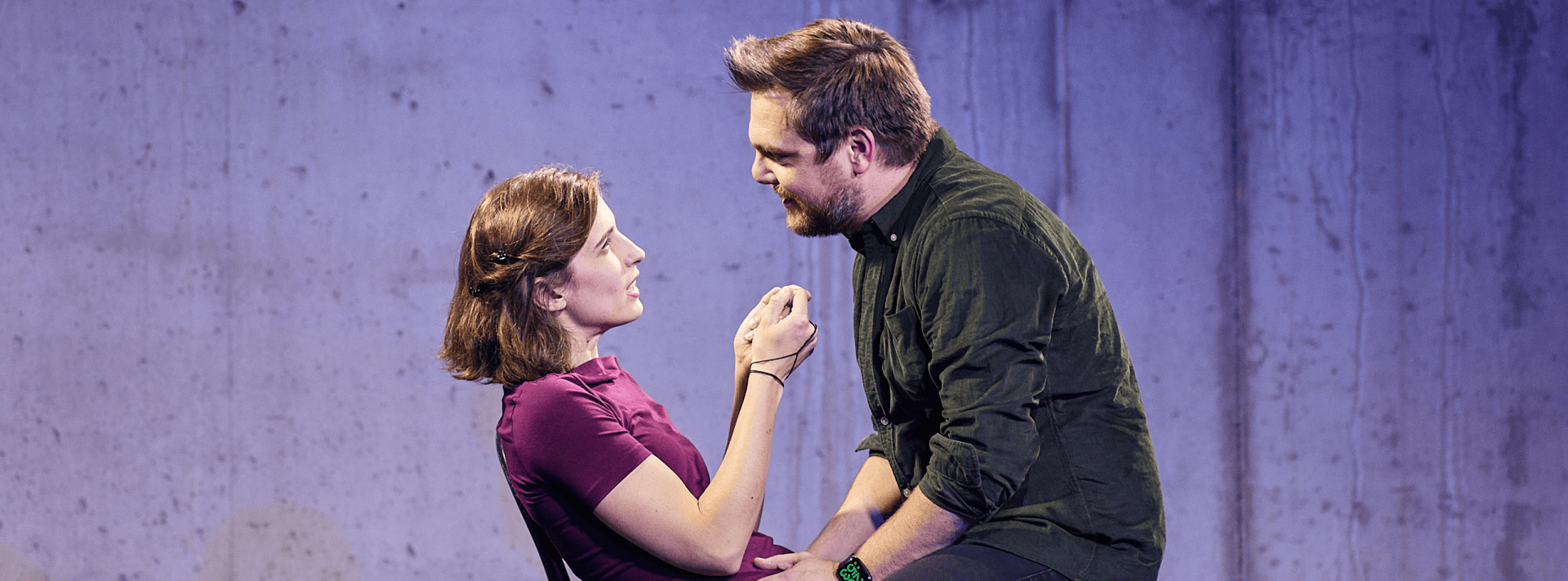 Eine Frau sitzt auf einem Stuhl gegenüber einem Mann, der auf einem Tisch sitzt und ihre Hand hält. Vor einem schlichten, hellvioletten Hintergrund blicken sie sich in die Augen.