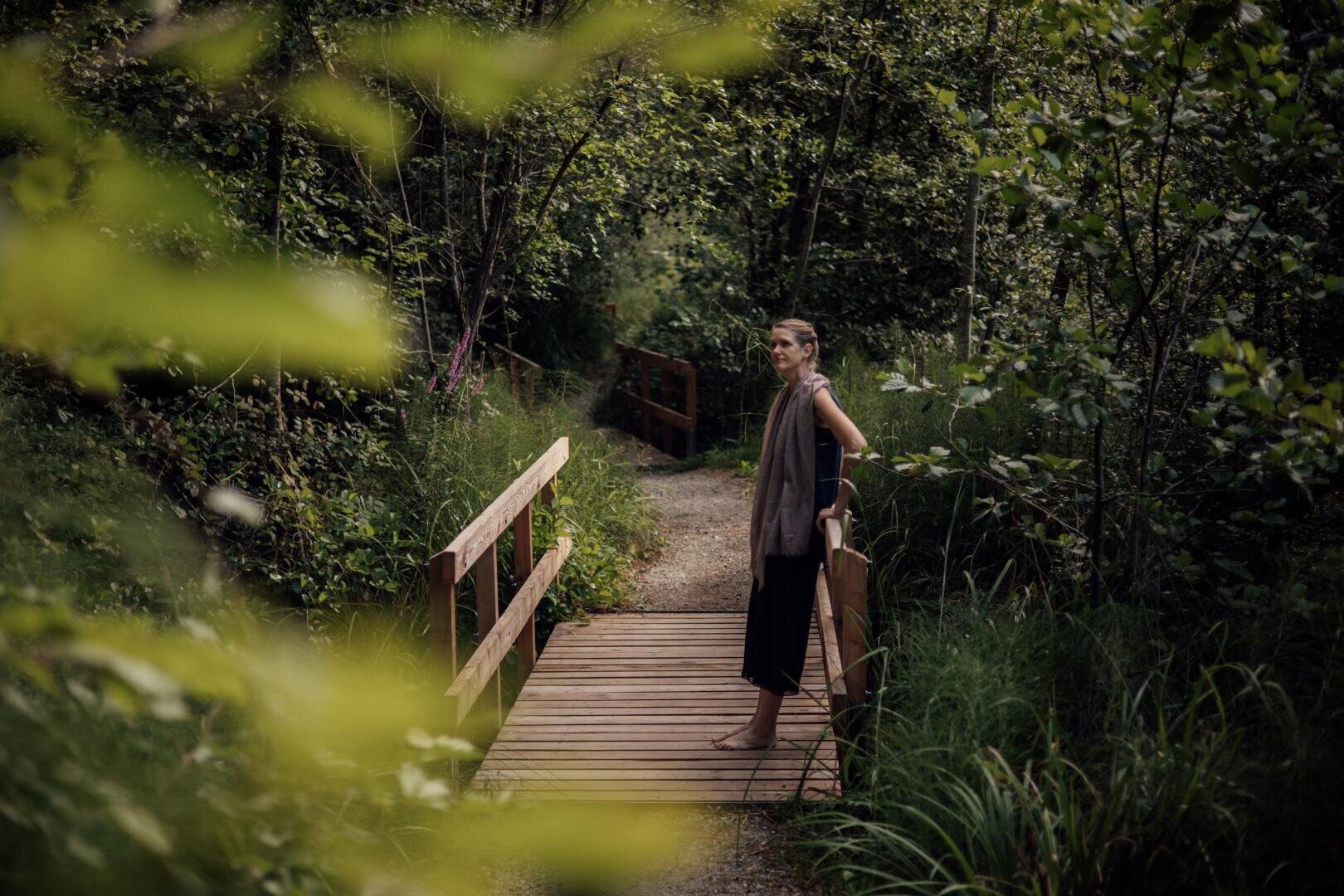 Eine Person steht barfuß auf einer Holzbrücke in einem üppig grünen Wald, umgeben von hohen Bäumen und Pflanzen, und blickt nachdenklich in die Ferne.