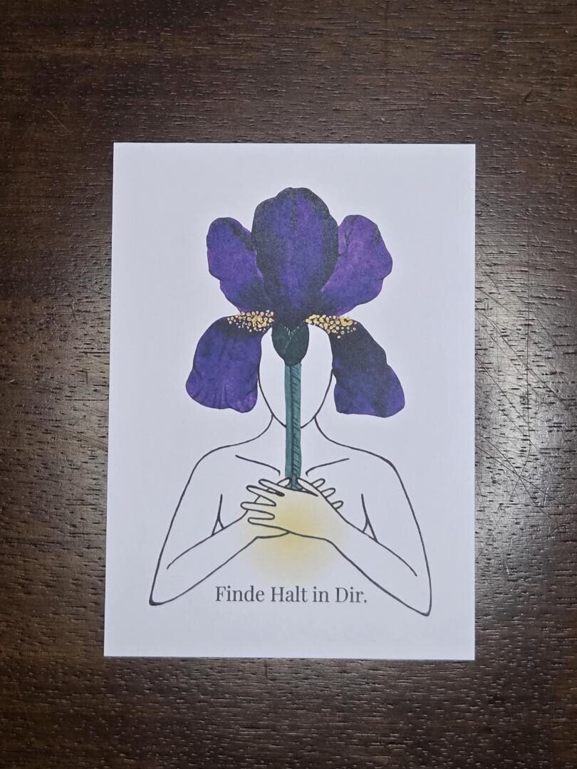 Eine künstlerisch gestaltete Karte mit einem menschlichen Oberkörper liegt auf hölzernem Hintergrund. Aus dem Herzen der Figur wächst eine violette Irisblüte, die deren Kopf ersetzt. Im Brustberiech leuchtet zudem ein gelber Lichtkreis. Darunter steht: "Finde Halt in dir".
