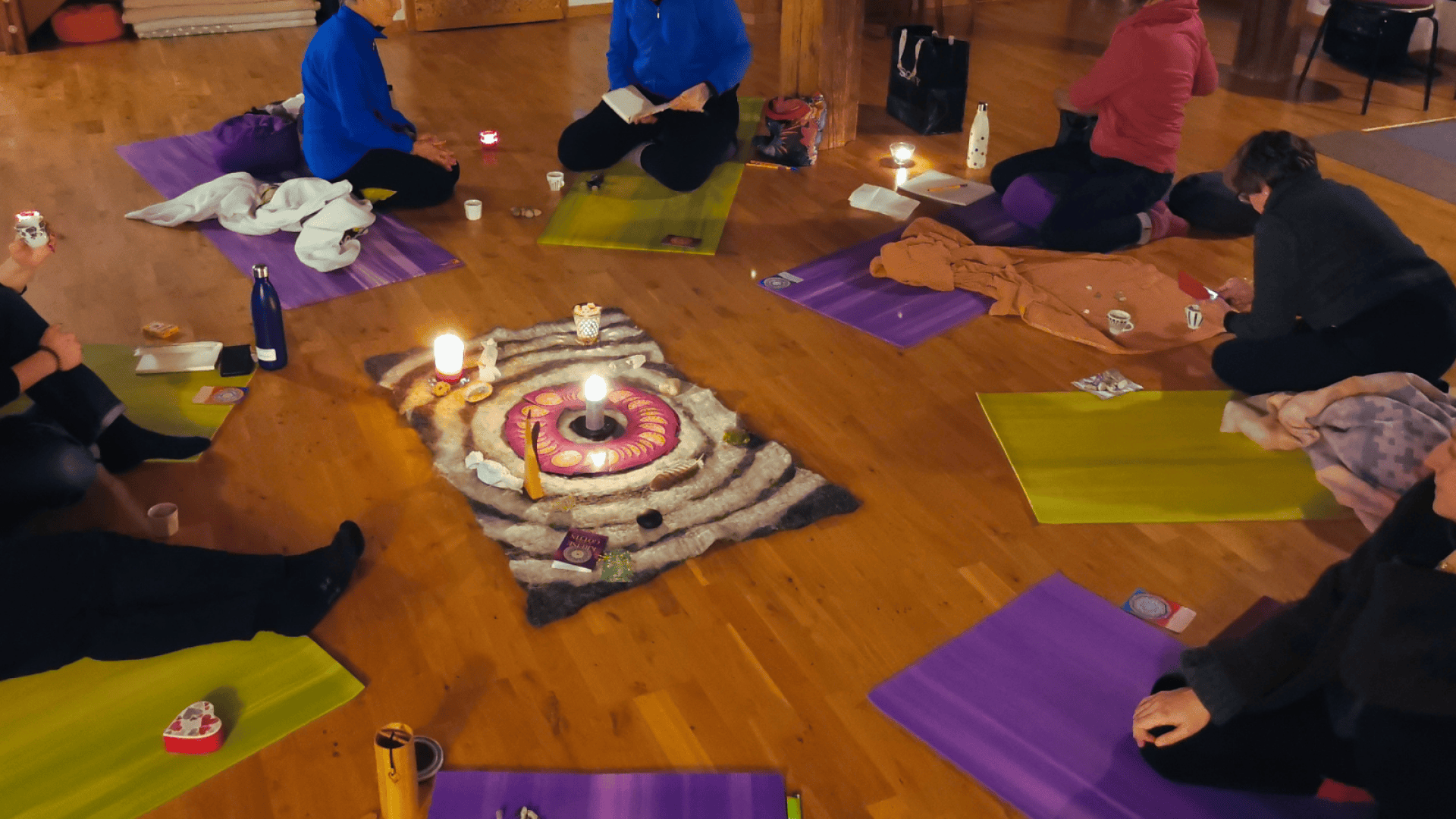 Eine Gruppe von Menschen sitzt auf Yogamatten im Kreis um einen Teppich in der Mitte, auf dem Kerzen und kleine Ritualgegenstände stehen, auf einem Holzboden.