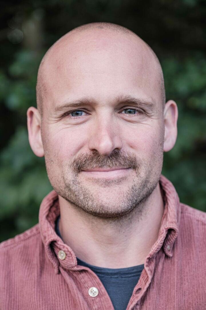 Ein Mann mit rasiertem Kopf, hellem Schnurrbart und Bart, der ein rötliches Kordhemd über einem dunklen T-Shirt trägt, steht im Freien, während das Grün im Hintergrund verschwimmt.