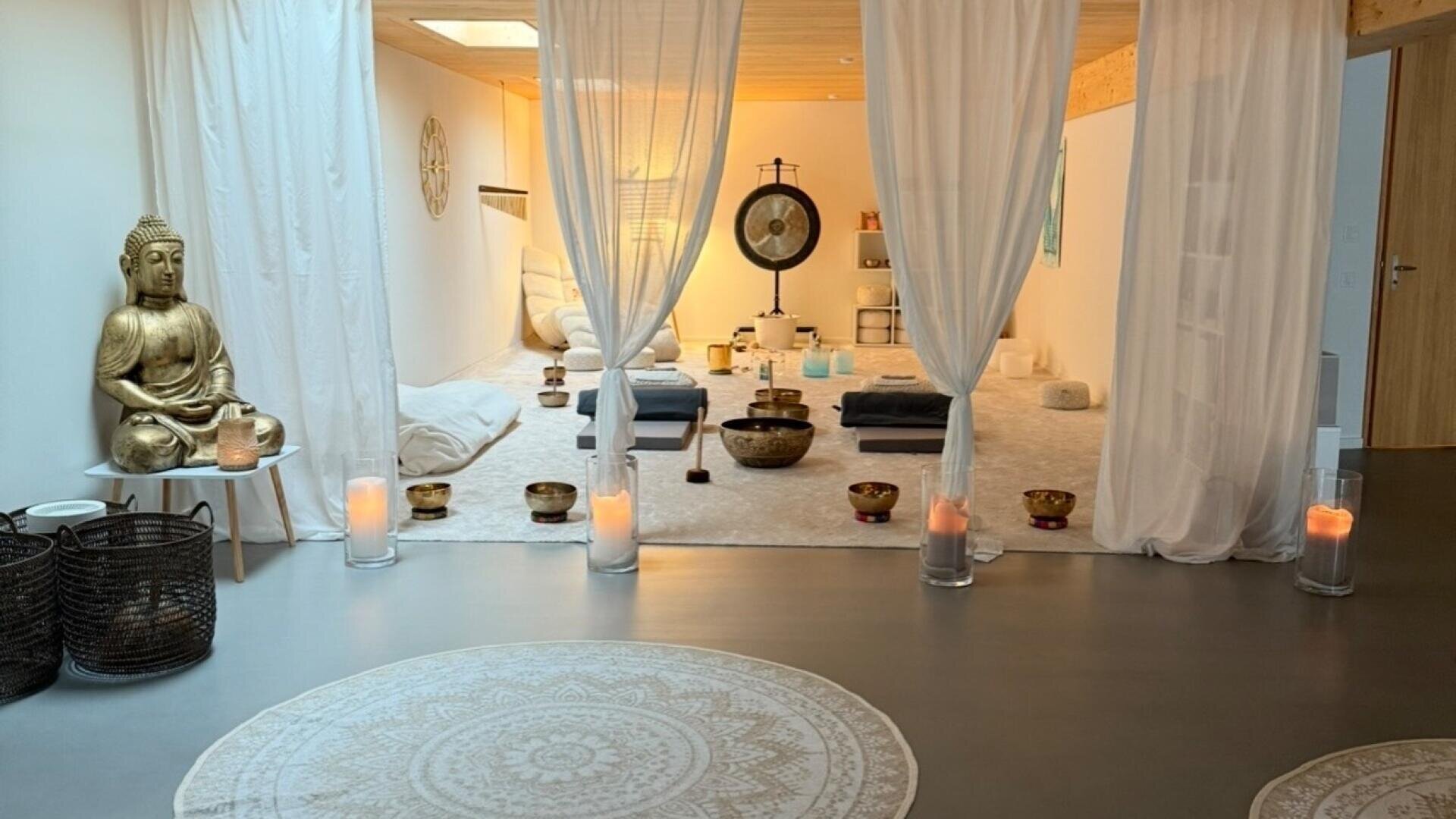 Ein ruhiger Meditationsraum mit runden gemusterten Teppichen, weißen Vorhängen, Kerzen, einer goldenen Buddha-Statue, Kissen und Klangschalen, die ordentlich angeordnet sind, alles in einer ruhigen, einladenden Atmosphäre.