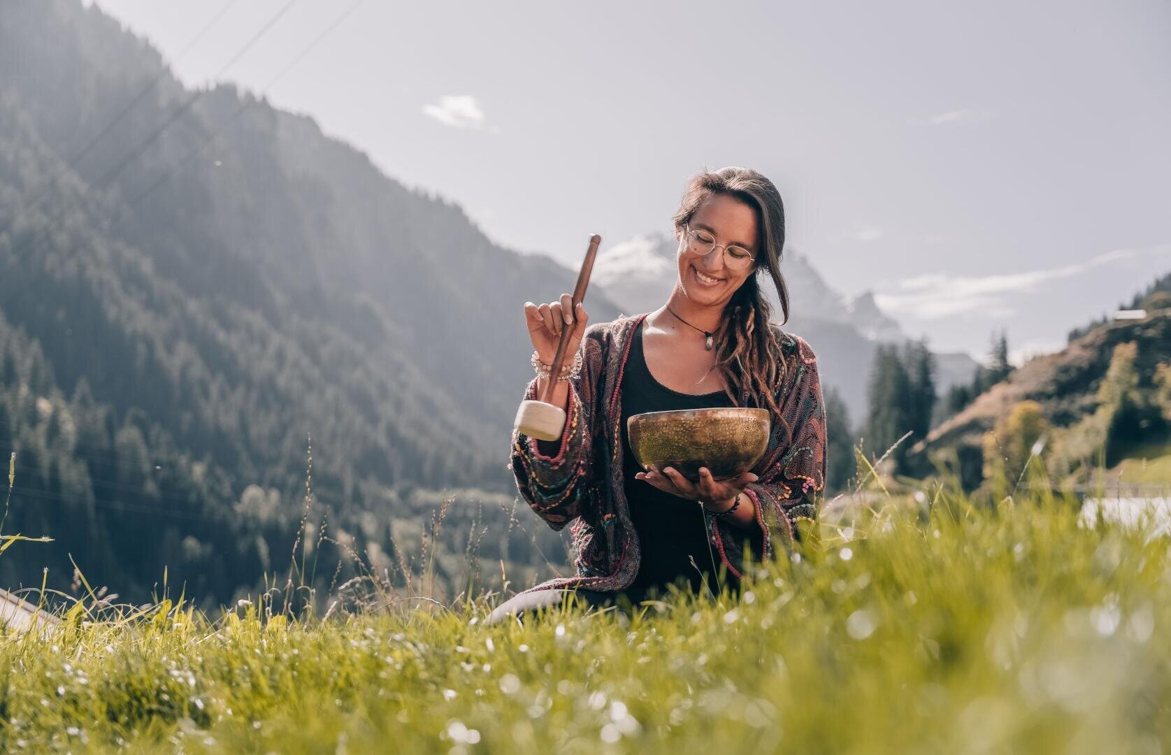 Eine Frau sitzt auf einer Wiese in den Bergen, lächelt und schlägt unter strahlend blauem Himmel mit einem Schlägel auf eine Klangschale.