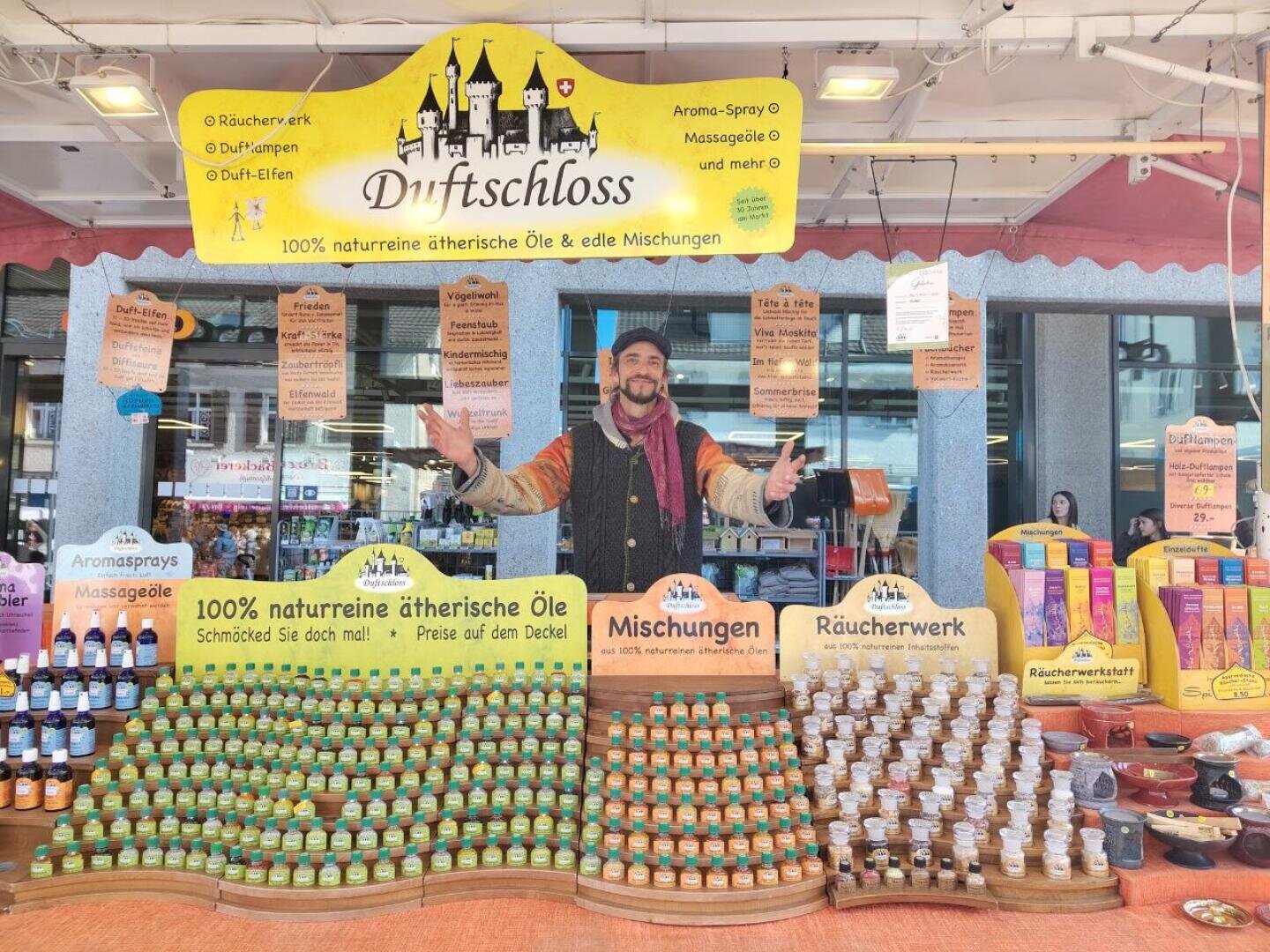 Ein fröhlicher Verkäufer steht hinter einem bunten Marktstand mit ätherischen Ölen, Sprays und Weihrauch. Auf den Schildern über dem Stand steht Duftschloss und wirbt für natürliche, aromatische Produkte. Flaschen und Tiegel stehen fein säuberlich auf dem Tisch.