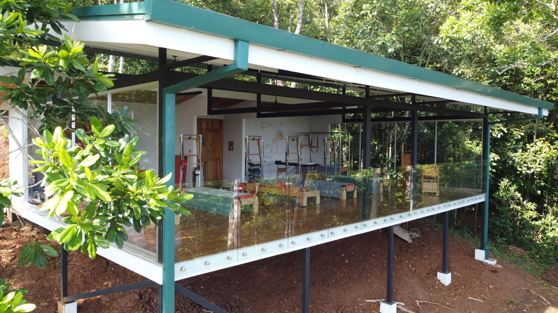 Ein modernes, offenes Klassenzimmer mit Glaswänden, Metallstützen und einem grünen Dach ist von einem üppigen Wald umgeben. Im Inneren sind Stühle und Staffeleien für den Kunstunterricht aufgestellt.