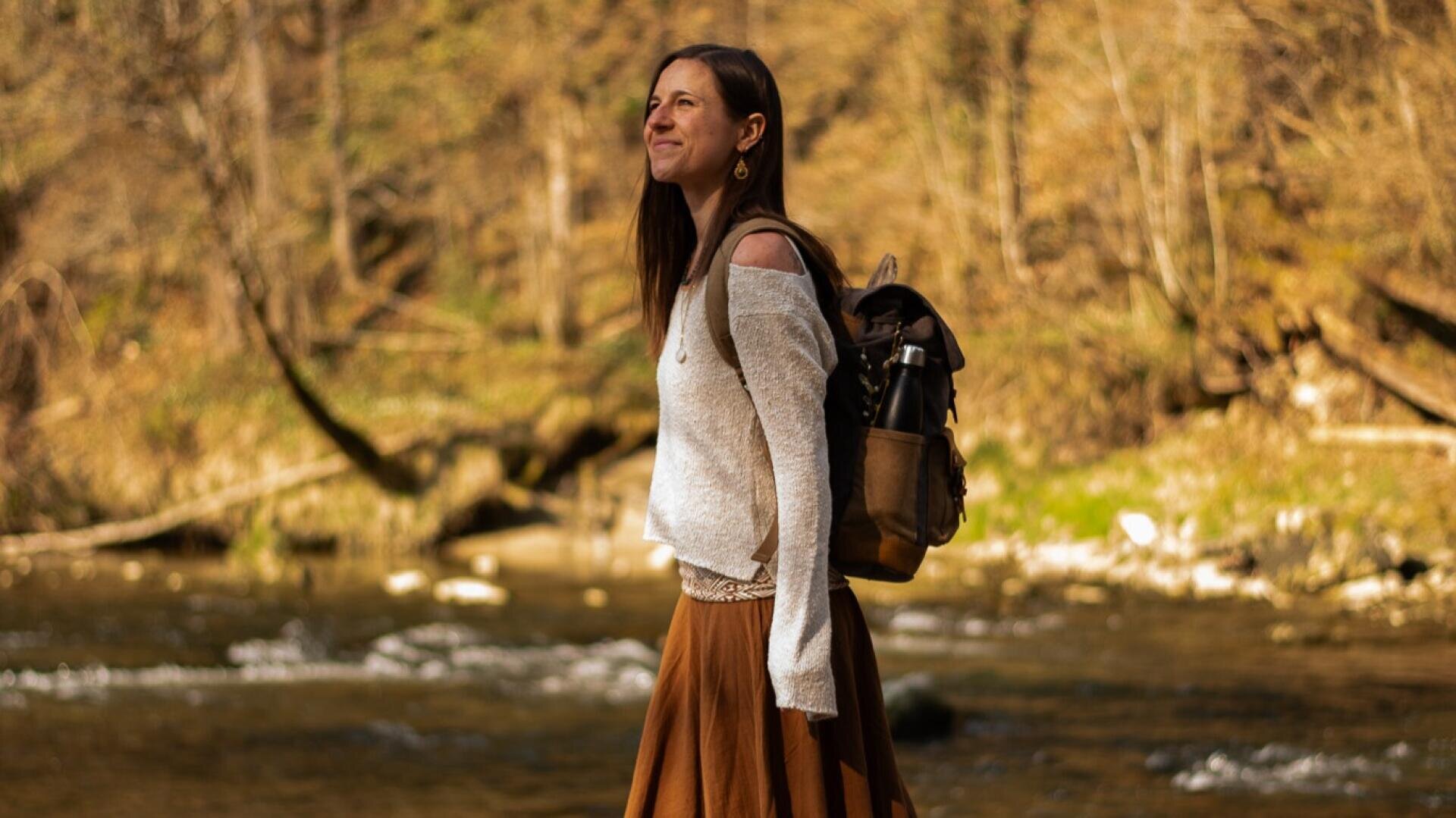 Eine Frau mit langen braunen Haaren, die einen weißen Pullover und einen braunen Rock trägt, steht im Freien an einem Fluss mit Bäumen im Hintergrund, lächelt und hält einen schwarzen Rucksack in der Hand.